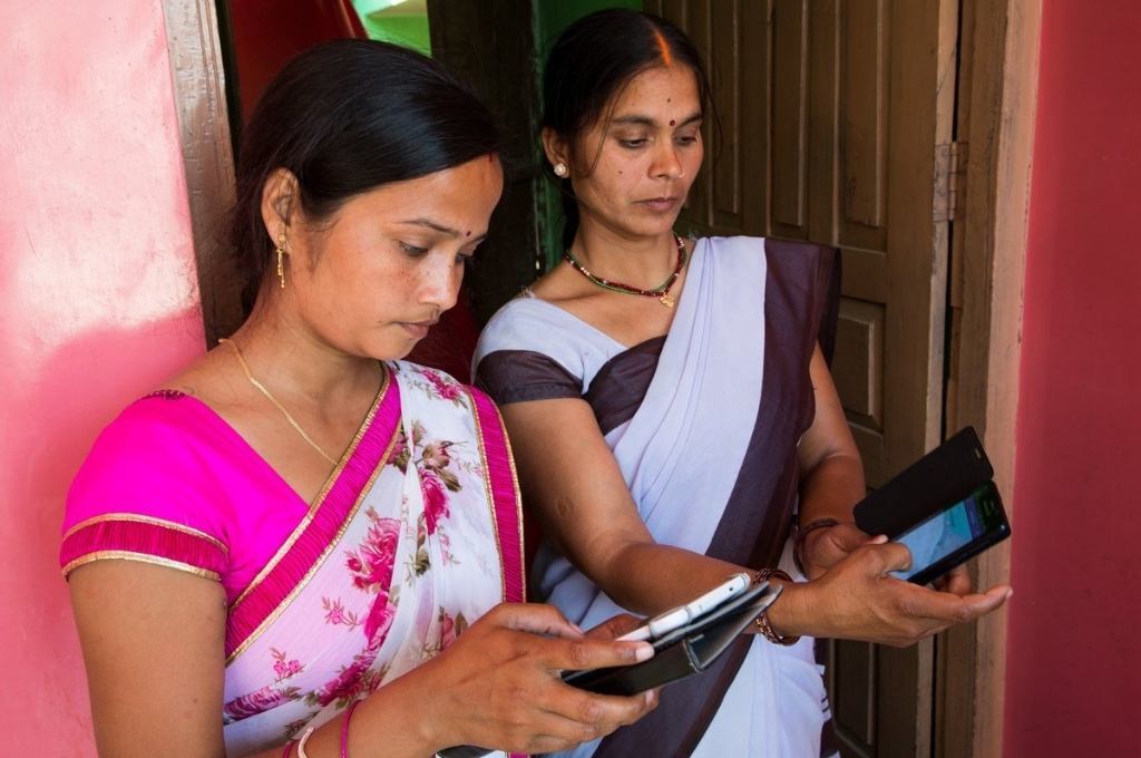 दो महिलाएं, एक अपने स्मार्टफोन को देख रही है और एक टैबलेट को देख रही है और ऐप का उपयोग कर रही है-ई-श्रम पोर्टल