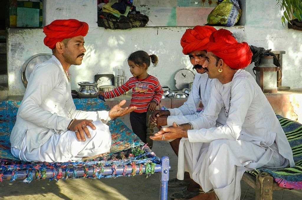 लाल पगड़ी और सफ़ेद कुर्ता पजामा पहने तीन लोग एक चारपाई पर बैठे हैं और उनके पीछे एक बच्ची खड़ी है-तलाक़ रबारी समुदाय राजस्थान