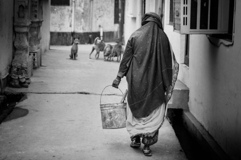 हाथ में बाल्टी लेकर सड़क पर चलती महिला पीछे से दिख रही है-दलित समुदाय