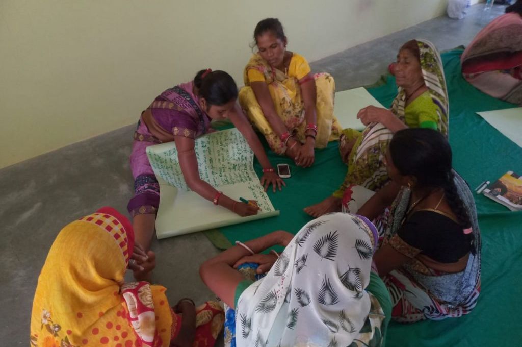 कोकिला महिलाओं के एक समूह में बैठकर एक रजिस्टर में कुछ लिखती हुई_आदिवासी महिला