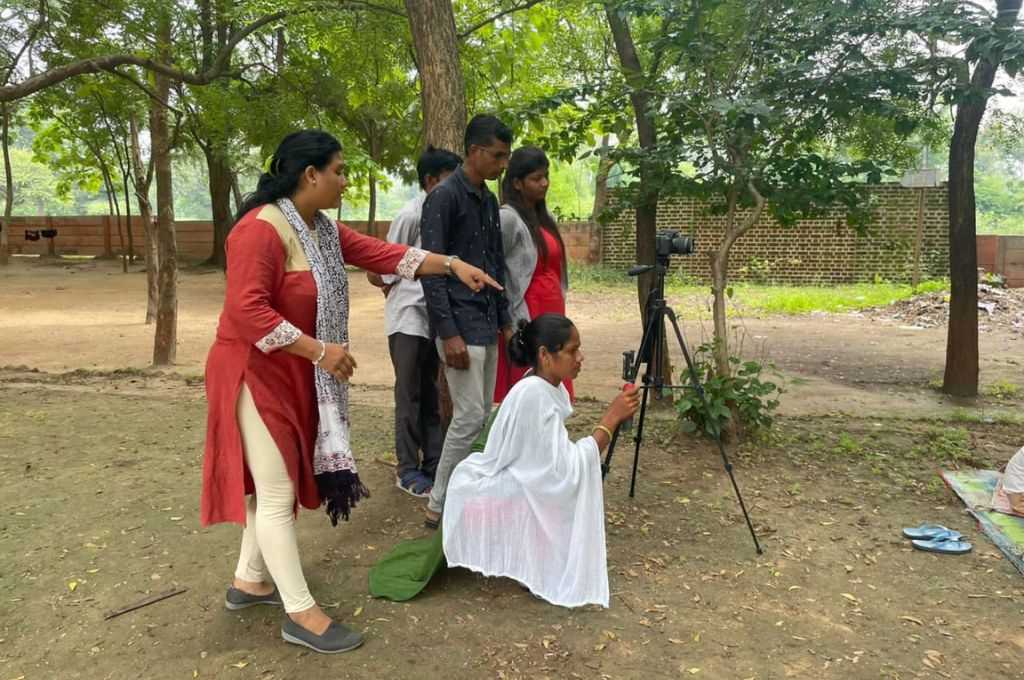 अपने आसपास के लोगों के साथ पार्क में कैमरे से शूटिंग करती एक महिला_आदिवासी पत्रकार