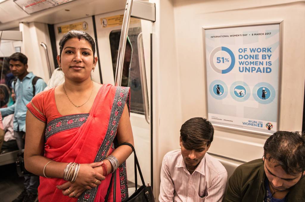 मेट्रो में खड़ी एक महिला। पीछे एक पोस्टर पर लिखा है ‘महिलाओं द्वारा किए गए 51 प्रतिशत काम का भुगतान नहीं किया जाता है’।