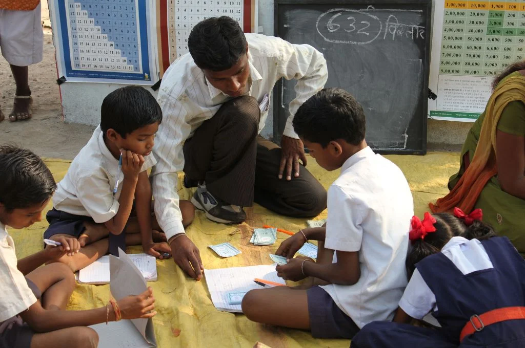 एक शिक्षक ज़मीन पर बैठकर दो छात्रों को पढ़ाता हुआ_ शिक्षक
