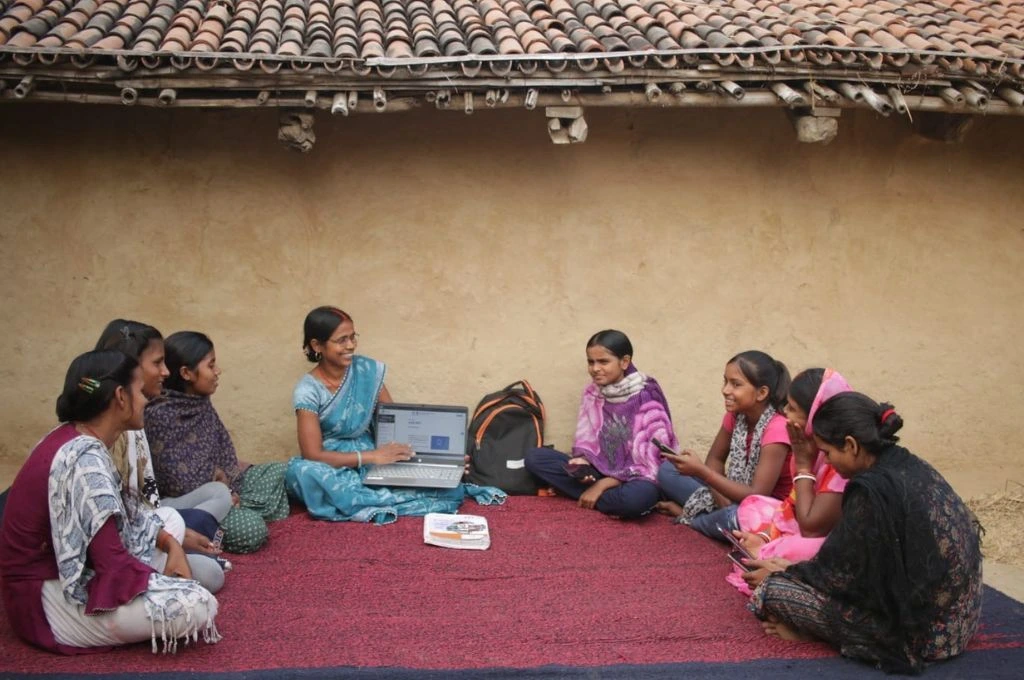 ग्रामीण आंगन में महिलाएं दरी पर बैठी हैं, एक महिला लैपटॉप के ज़रिए जानकारी दे रही हैं_आर्टिफिशियल इंटेलिजेंस
