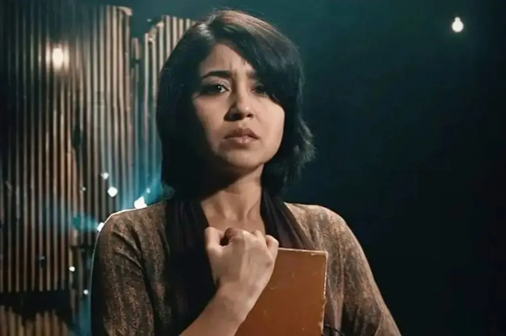एक युवती गंभीर भाव-भंगिमा के साथ सामने देख रही है। वह दोनों हाथों से एक किताब को अपने सीने से लगाए हुए है। बैकग्राउंड में हल्की रोशनी और पर्दे जैसे स्ट्रक्चर हैं, जो मंच या स्टूडियो का आभास देते हैं_फील्ड ट्रिप