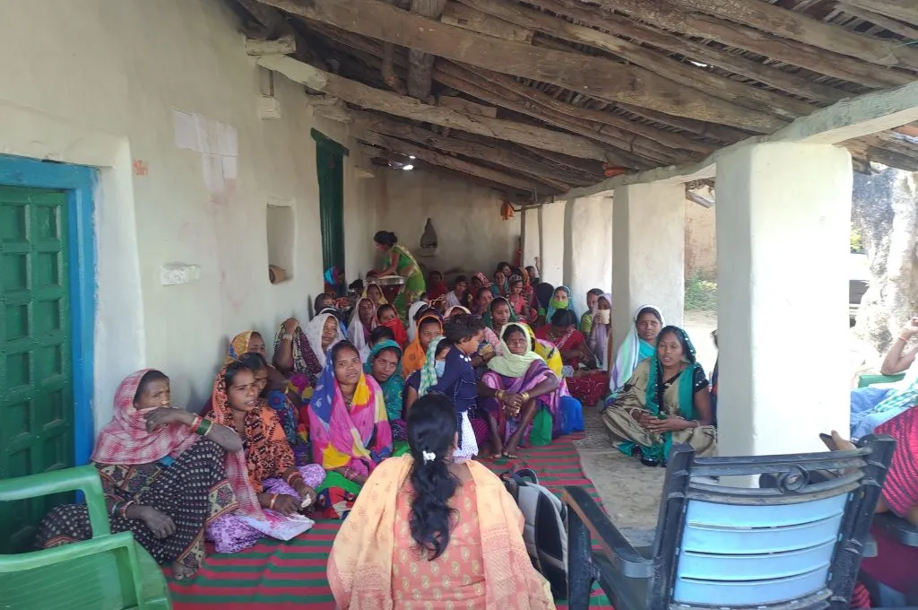 धरती पर बैठे महिलाओं के समूह को संबोधित करती एक महिला_संगठन