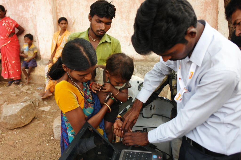 एक महिला गोद में बच्चे को लिए है, और एक व्यक्ति उसका हाथ पकड़कर किसी मशीन से जाँच कर रहा है_डिजिटल वेलफेयर