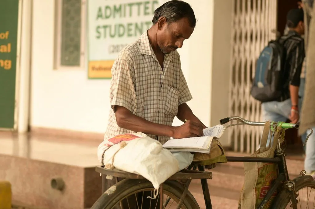 साइकिल की गद्दी पर कॉपी में कुछ लिखते हुए एक व्यक्ति_युवा श्रम बल