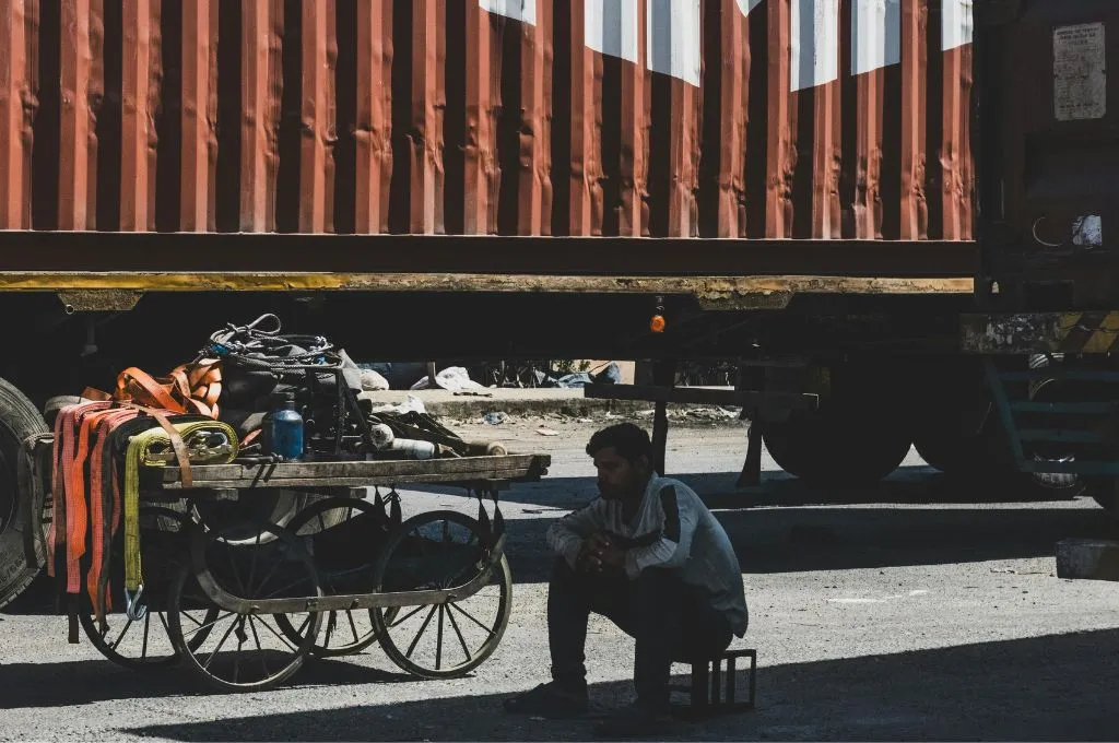 सड़क किनारे खड़े ट्रक और रेहड़े के बराबर में छोटे स्टूल पर आराम की मुद्रा में बैठा एक व्यक्ति_युवा श्रम बल