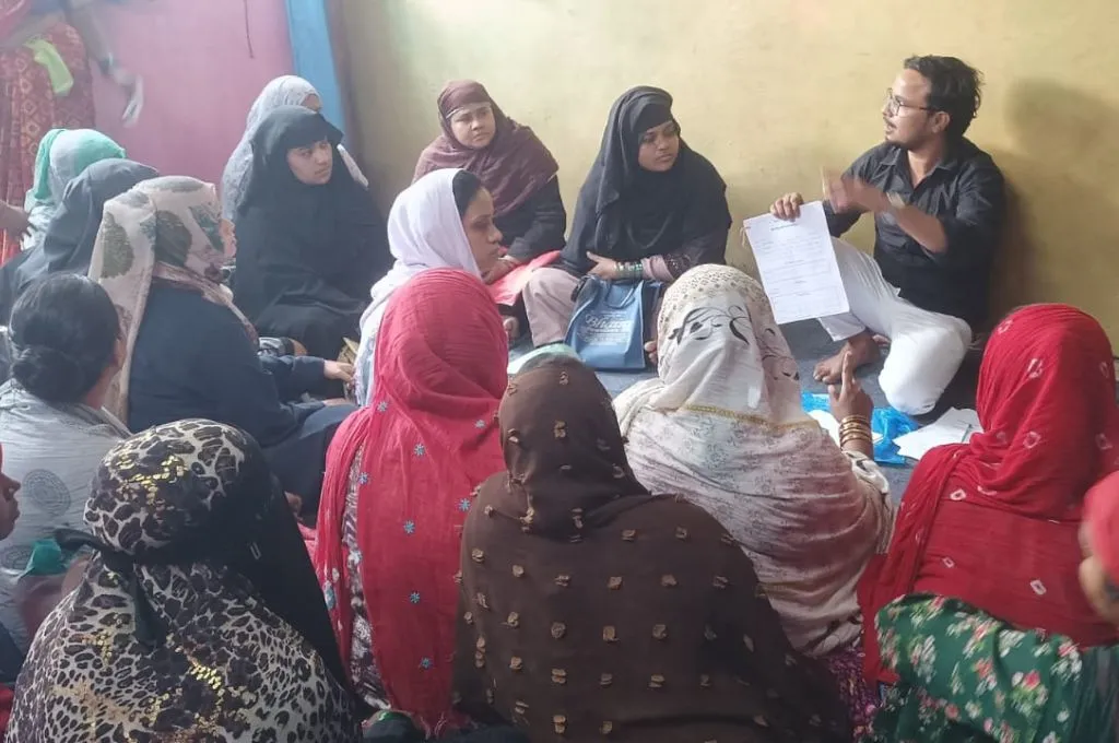 जमीन पर घेरे में बैठी महिलाओं के समूह को संबोधित करता उन्हीं के साथ बैठा एक पुरुष_प्रवासी श्रमिक