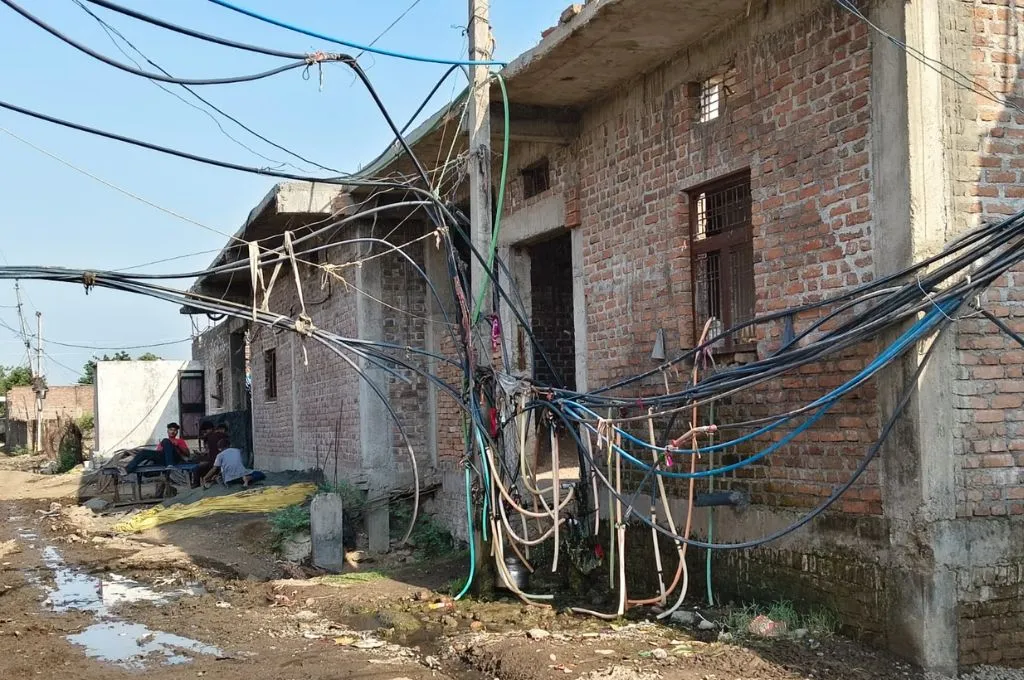 ईंट से बने घरों के सामने एक टूटी सड़क और बिजली के तारों का जाल और खटिया पर बैठे कुछ लोग_ छबड़ा थर्मल पावर प्लांट