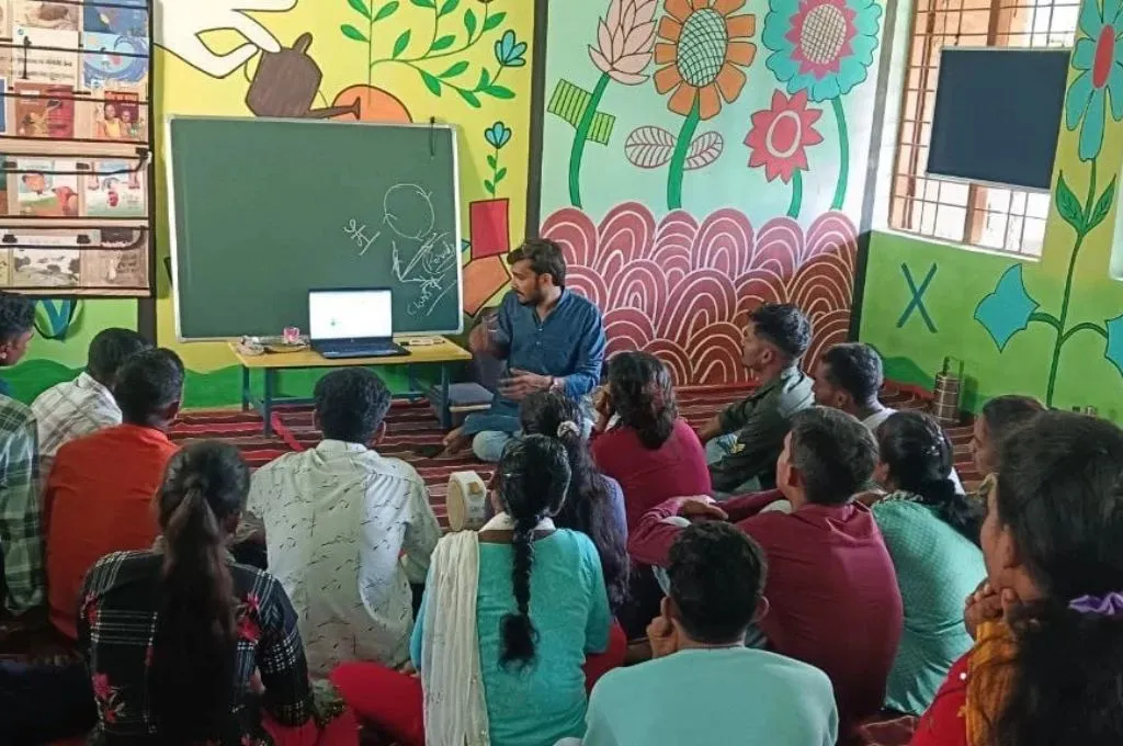 एक कमरे में ज़मीन पर बैठकर एक वक्ता को सुनते हुए कुछ लोग_मानसिक स्वास्थ्य